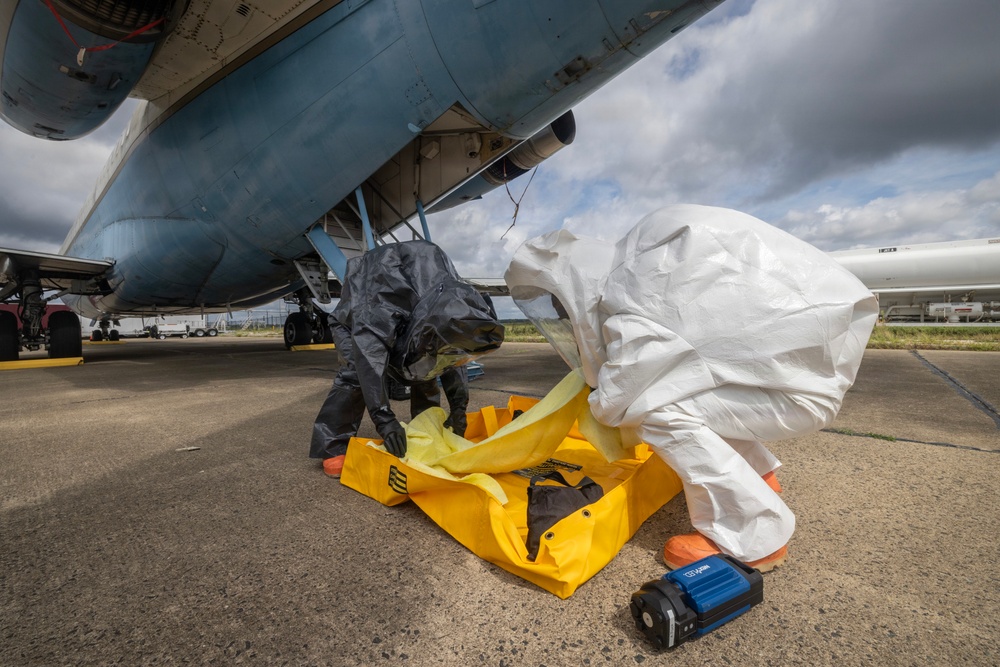 Two personnel in full hazmat/chemical protective suits are working beneath the fuselage of a large military or government aircraft on a tarmac. They appear to be handling or deploying yellow containment equipment, likely as part of a HAZMAT response drill or actual incident response. A blue electronic detection device is visible on the ground nearby.