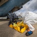Two personnel in full hazmat/chemical protective suits are working beneath the fuselage of a large military or government aircraft on a tarmac. They appear to be handling or deploying yellow containment equipment, likely as part of a HAZMAT response drill or actual incident response. A blue electronic detection device is visible on the ground nearby.