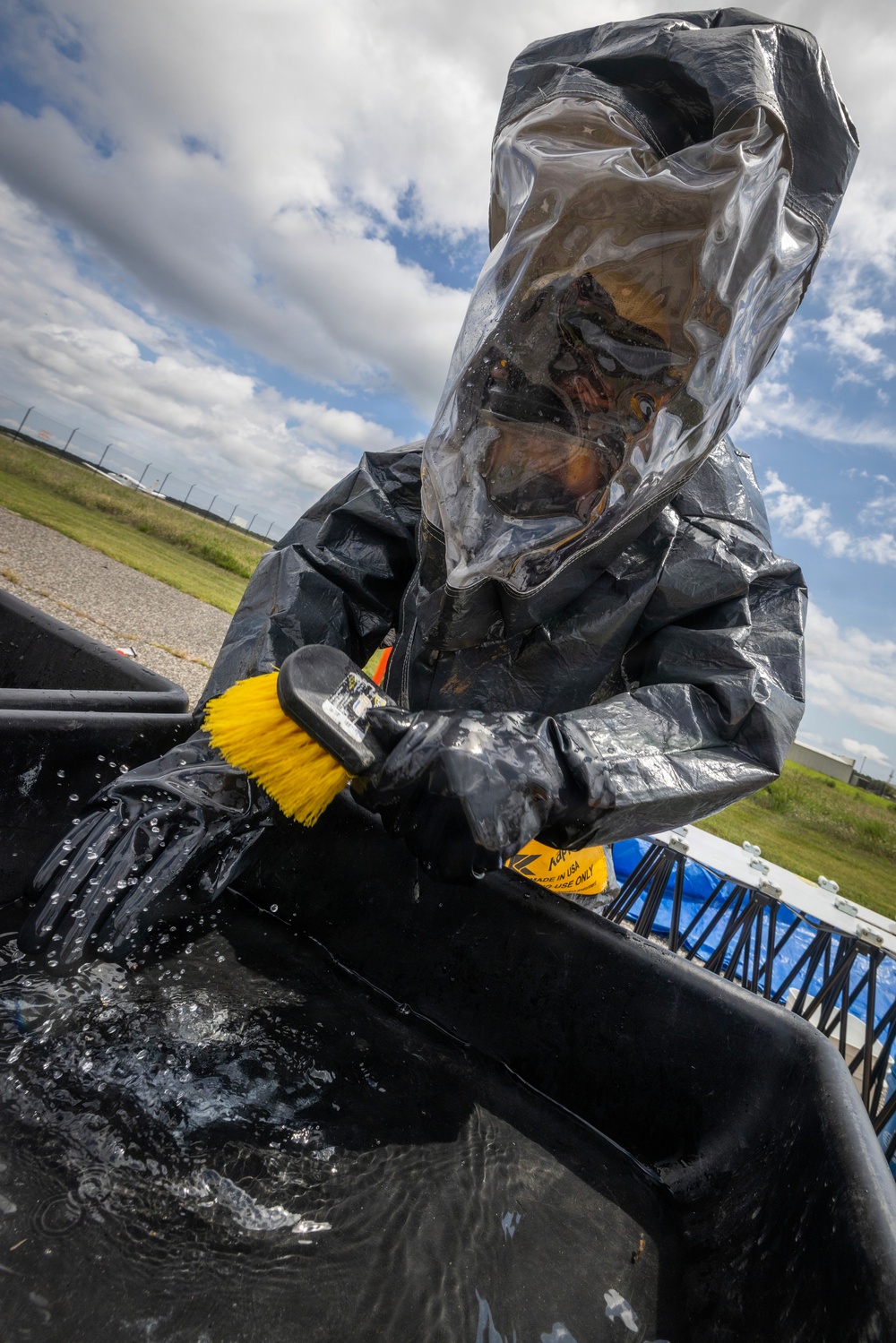 A person wearing full hazmat/chemical protective gear, including a black encapsulated suit with a clear transparent hood and rubber gloves, is scrubbing or cleaning a surface using a yellow-bristled brush. The individual is leaning over what appears to be a black containment vessel or trough filled with water. The setting appears to be an outdoor area, likely a military base or industrial facility, under a partly cloudy sky.