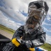 A person wearing full hazmat/chemical protective gear, including a black encapsulated suit with a clear transparent hood and rubber gloves, is scrubbing or cleaning a surface using a yellow-bristled brush. The individual is leaning over what appears to be a black containment vessel or trough filled with water. The setting appears to be an outdoor area, likely a military base or industrial facility, under a partly cloudy sky.