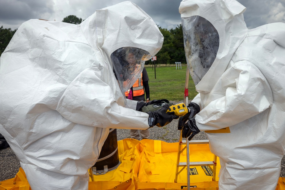 Two individuals wearing white full-body hazmat suits and black rubber gloves are handling a yellow handheld detection device, likely a multi-gas or radiation detector. They appear to be conducting a hazardous materials (HAZMAT) response exercise or operation, with a yellow containment berm visible in the foreground. A third person in an orange safety vest observes in the background.