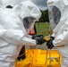 Two individuals wearing white full-body hazmat suits and black rubber gloves are handling a yellow handheld detection device, likely a multi-gas or radiation detector. They appear to be conducting a hazardous materials (HAZMAT) response exercise or operation, with a yellow containment berm visible in the foreground. A third person in an orange safety vest observes in the background.