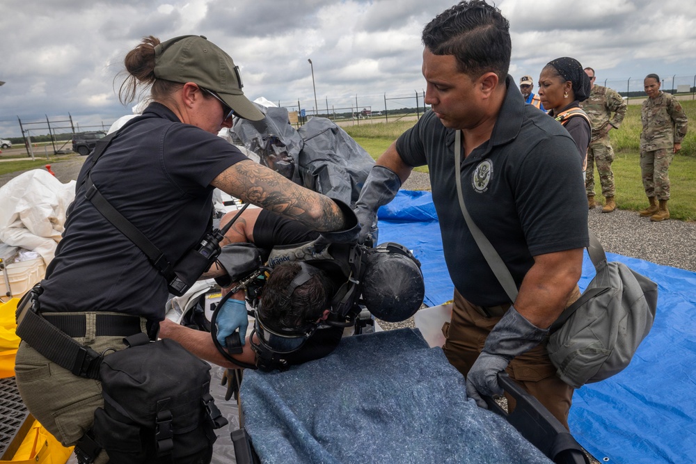 Two personnel in dark tactical/law enforcement attire wearing blue nitrile gloves are attending to a person lying on a blue tarp-covered surface, appearing to remove or adjust protective gear including a helmet. Military personnel in camouflage uniforms observe in the background near what appears to be an airfield or tarmac. The scene resembles a training exercise or emergency response drill.