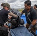 Two personnel in dark tactical/law enforcement attire wearing blue nitrile gloves are attending to a person lying on a blue tarp-covered surface, appearing to remove or adjust protective gear including a helmet. Military personnel in camouflage uniforms observe in the background near what appears to be an airfield or tarmac. The scene resembles a training exercise or emergency response drill.