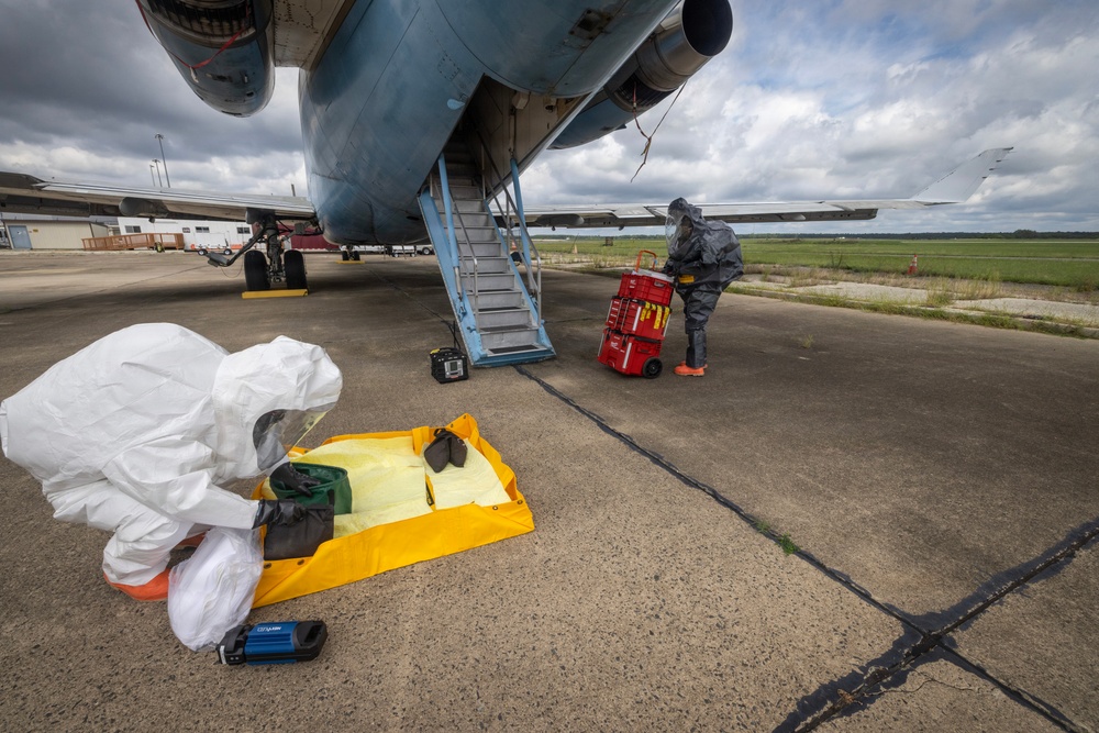 Two individuals wearing full hazmat/CBRN protective suits are conducting operations on an airport tarmac next to a large military or cargo aircraft. One person is crouched over a yellow containment mat with equipment laid out, while the second person is handling a red wheeled equipment cart near the aircraft's boarding stairs. The scene appears to be a hazardous materials response exercise or actual HAZMAT operation.