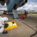 Two individuals wearing full hazmat/CBRN protective suits are conducting operations on an airport tarmac next to a large military or cargo aircraft. One person is crouched over a yellow containment mat with equipment laid out, while the second person is handling a red wheeled equipment cart near the aircraft's boarding stairs. The scene appears to be a hazardous materials response exercise or actual HAZMAT operation.