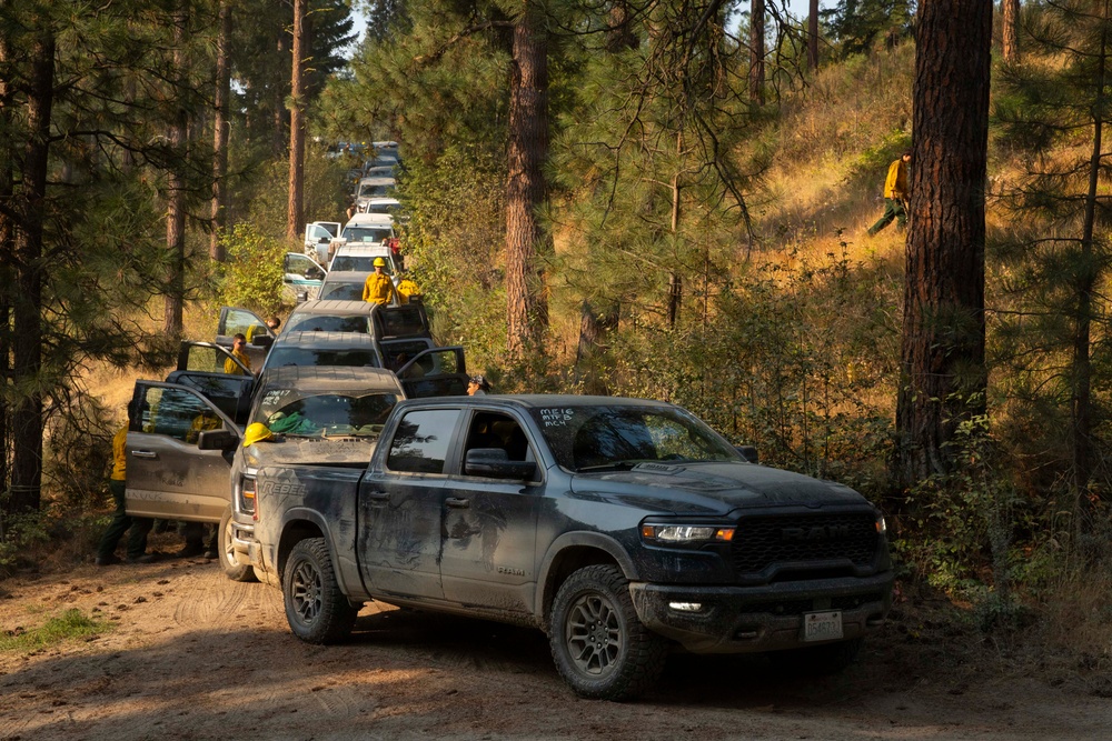 A convoy of pickup trucks and vehicles is lined up on a dirt forest road, with personnel wearing yellow fire-resistant gear visible alongside and on the vehicles. The scene depicts what appears to be a wildfire response operation, with firefighters or forestry workers mobilizing through a pine forest. The lead vehicle is a dark gray Ram Rebel pickup truck, followed by several other trucks stretching into the background.