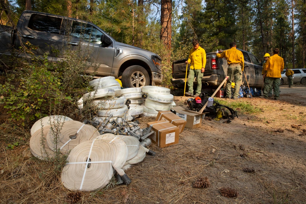 A group of wildland firefighters in yellow Nomex shirts are staged at a forest road location with two pickup trucks. In the foreground, a large cache of firefighting supplies is laid out on the ground, including rolled fire hoses, hose fittings/couplings, and cardboard supply boxes. The setting appears to be a conifer forest, likely during an active wildfire response operation.