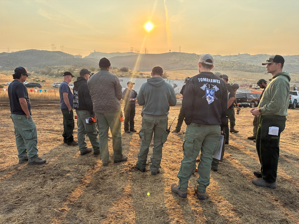 A group of approximately 10-15 personnel are gathered in a circle for what appears to be a briefing or debriefing session in an outdoor field setting. One individual is wearing a jacket with 'TOMAHAWKS' and a shield emblem visible on the back, suggesting a firefighting crew or tactical team. The scene is set against a hazy, smoke-filtered sunset over rolling hills, consistent with a wildfire incident command environment.
