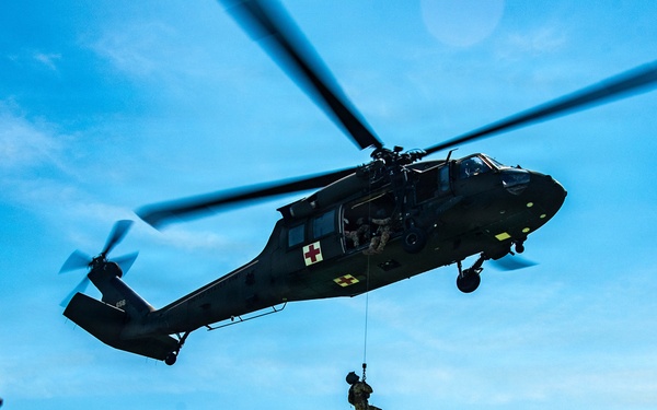 Two military personnel in camouflage uniforms observe a medical evacuation (MEDEVAC) helicopter hovering overhead while a soldier is being hoisted on a cable below the aircraft. The helicopter displays red cross markings on a yellow background, identifying it as a medical/MEDEVAC variant of the UH-60 Black Hawk. The scene appears to be a training exercise or demonstration conducted in a semi-urban environment.