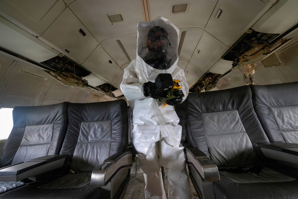 A person wearing a full white hazmat/protective suit with an enclosed hood and respirator stands in the aisle of an aircraft cabin. The individual is holding a yellow and black handheld device, possibly a detection instrument or sprayer. The aircraft interior shows signs of wear and partial disassembly, suggesting a decommissioned or training aircraft.