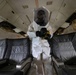 A person wearing a full white hazmat/protective suit with an enclosed hood and respirator stands in the aisle of an aircraft cabin. The individual is holding a yellow and black handheld device, possibly a detection instrument or sprayer. The aircraft interior shows signs of wear and partial disassembly, suggesting a decommissioned or training aircraft.