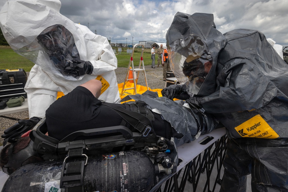 Two personnel in full hazmat/CBRN protective suits are attending to a person lying on a stretcher or table, appearing to conduct a decontamination or medical response exercise. One responder wears a white Tyvek-style suit and the other wears a dark gray encapsulated suit, both bearing 'Kappler' brand labels. In the background, additional personnel in military and civilian attire observe the exercise at what appears to be an outdoor training facility.