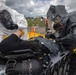 Two personnel in full hazmat/CBRN protective suits are attending to a person lying on a stretcher or table, appearing to conduct a decontamination or medical response exercise. One responder wears a white Tyvek-style suit and the other wears a dark gray encapsulated suit, both bearing 'Kappler' brand labels. In the background, additional personnel in military and civilian attire observe the exercise at what appears to be an outdoor training facility.