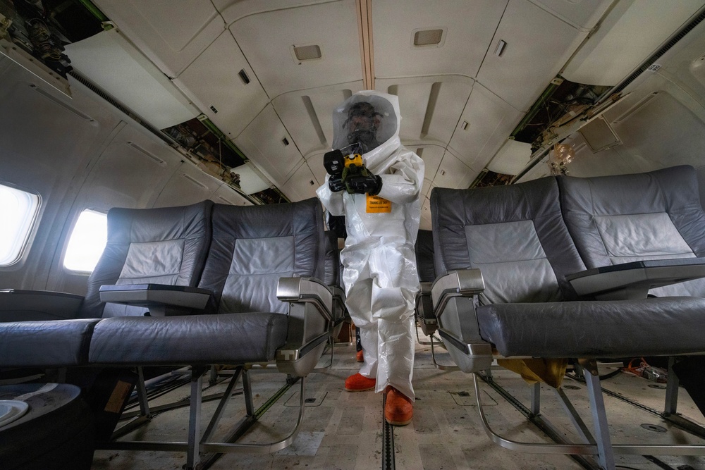 A person wearing a full white hazmat/protective suit with a clear face shield and orange boot covers stands in the aisle of a commercial aircraft cabin. They are holding a yellow handheld device, likely a scanner or sprayer, suggesting decontamination or inspection work. The aircraft interior appears worn or partially dismantled, with overhead panels showing exposed components.