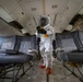 A person wearing a full white hazmat/protective suit with a clear face shield and orange boot covers stands in the aisle of a commercial aircraft cabin. They are holding a yellow handheld device, likely a scanner or sprayer, suggesting decontamination or inspection work. The aircraft interior appears worn or partially dismantled, with overhead panels showing exposed components.