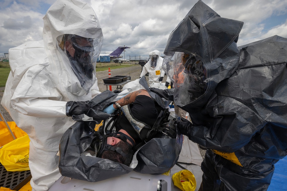 A HAZMAT decontamination exercise or response operation is underway outdoors, with personnel in full protective suits attending to a person lying on a stretcher or decontamination surface. Two responders in different colored hazmat suits (white and dark gray/black) are actively treating or decontaminating the individual, who appears to have a breathing mask applied. A third responder in a white suit is visible in the background near decontamination equipment.