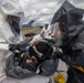 A HAZMAT decontamination exercise or response operation is underway outdoors, with personnel in full protective suits attending to a person lying on a stretcher or decontamination surface. Two responders in different colored hazmat suits (white and dark gray/black) are actively treating or decontaminating the individual, who appears to have a breathing mask applied. A third responder in a white suit is visible in the background near decontamination equipment.