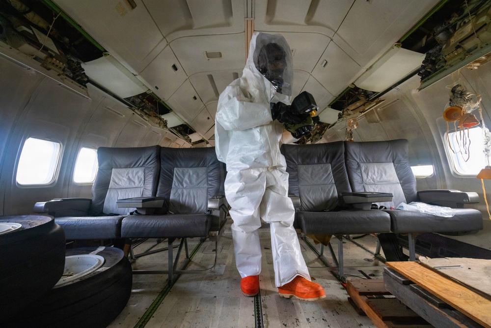 A person wearing a full white hazmat/Tyvek protective suit, gas mask, and orange rubber boots stands inside the deteriorated cabin of a decommissioned or damaged aircraft. The interior shows significant wear and damage, with exposed ceiling panels and missing overhead compartment covers. The individual appears to be conducting an inspection, decontamination, or forensic documentation activity.