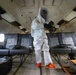 A person wearing a full white hazmat/Tyvek protective suit, gas mask, and orange rubber boots stands inside the deteriorated cabin of a decommissioned or damaged aircraft. The interior shows significant wear and damage, with exposed ceiling panels and missing overhead compartment covers. The individual appears to be conducting an inspection, decontamination, or forensic documentation activity.