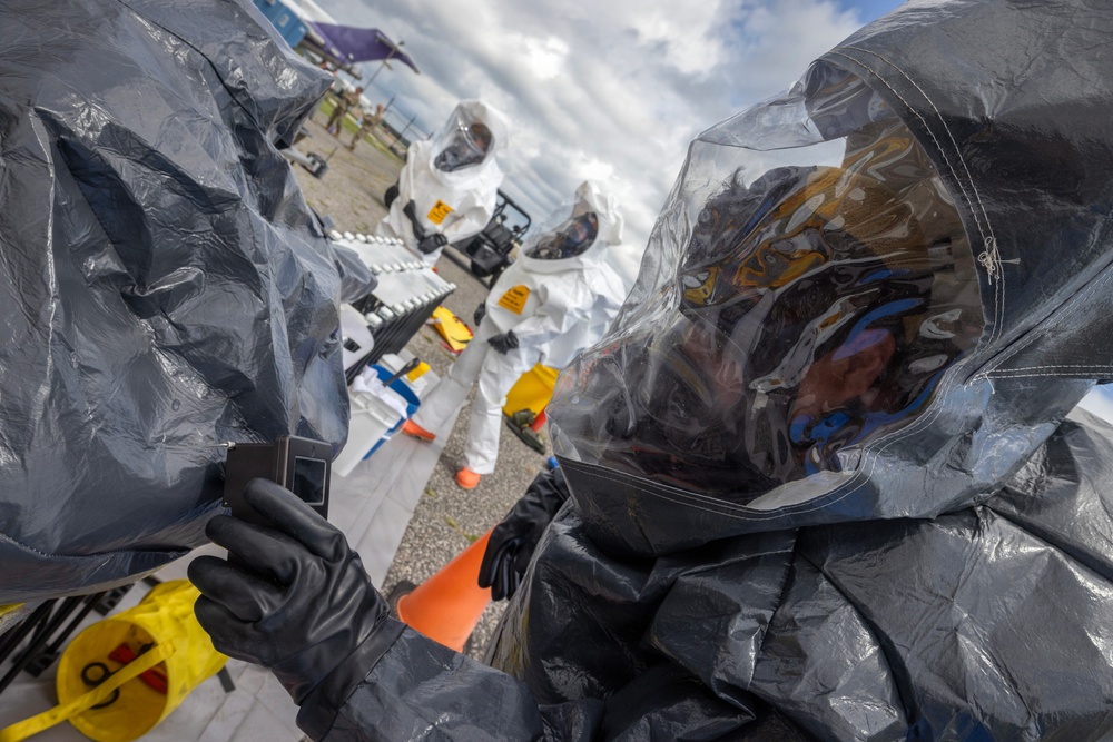 Personnel in full hazardous materials (HAZMAT) protective suits are engaged in what appears to be a CBRN (Chemical, Biological, Radiological, Nuclear) response exercise or operation. The foreground shows an individual in a dark gray encapsulated suit with a clear visor, while background personnel wear white Tyvek-style suits with respirators. Yellow hazard/radiation warning symbols are visible, suggesting a radiological or chemical incident scenario.