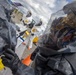 Personnel in full hazardous materials (HAZMAT) protective suits are engaged in what appears to be a CBRN (Chemical, Biological, Radiological, Nuclear) response exercise or operation. The foreground shows an individual in a dark gray encapsulated suit with a clear visor, while background personnel wear white Tyvek-style suits with respirators. Yellow hazard/radiation warning symbols are visible, suggesting a radiological or chemical incident scenario.
