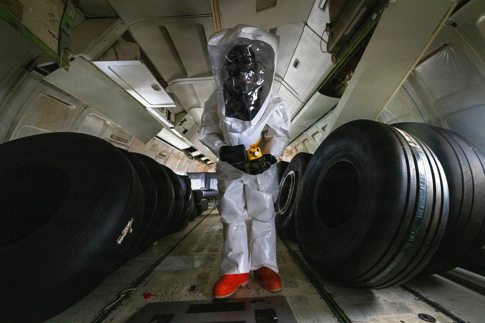 A person wearing a full white hazmat/CBRN protective suit with a clear plastic hood, orange boots, and black gloves is standing in what appears to be the cargo hold of a large aircraft. The individual is holding a yellow handheld detection instrument, likely a radiation or chemical detector. The cargo hold is loaded with large black aircraft tires arranged in rows on both sides.