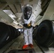 A person wearing a full white hazmat/CBRN protective suit with a clear plastic hood, orange boots, and black gloves is standing in what appears to be the cargo hold of a large aircraft. The individual is holding a yellow handheld detection instrument, likely a radiation or chemical detector. The cargo hold is loaded with large black aircraft tires arranged in rows on both sides.