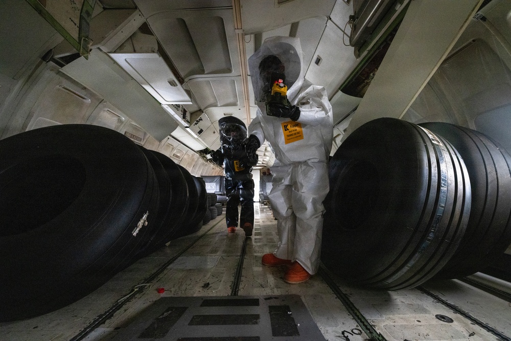 Two individuals wearing full hazmat/CBRN protective suits are working inside what appears to be the cargo hold of a large military or transport aircraft. One person wears a white Tyvek-style suit with orange boots and a transparent hood, while the other wears a black encapsulated suit. Both are carrying handheld detection or monitoring devices, suggesting a hazardous materials inspection or training exercise.