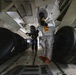 Two individuals wearing full hazmat/CBRN protective suits are working inside what appears to be the cargo hold of a large military or transport aircraft. One person wears a white Tyvek-style suit with orange boots and a transparent hood, while the other wears a black encapsulated suit. Both are carrying handheld detection or monitoring devices, suggesting a hazardous materials inspection or training exercise.