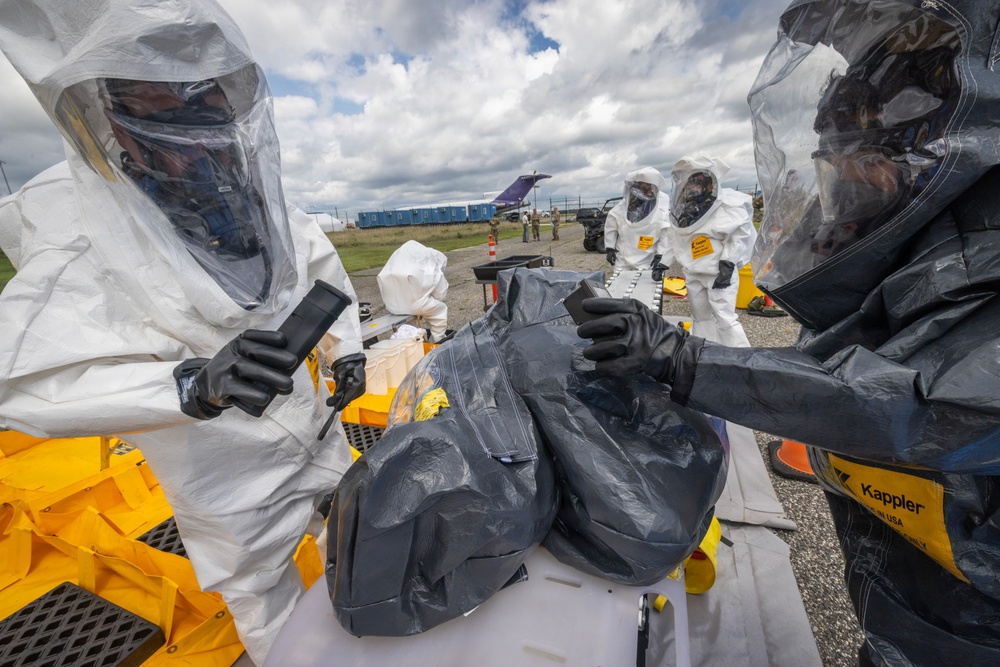 Personnel in full hazmat/CBRN protective suits are conducting what appears to be a decontamination or hazardous materials handling exercise at an outdoor staging area near an airfield. At least four individuals in white Tyvek-style suits with respirators and black gloves are working around a table with yellow equipment containers. The setting, equipment branding (Kappler), and organized staging suggest a military or emergency response training exercise.