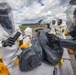 Personnel in full hazmat/CBRN protective suits are conducting what appears to be a decontamination or hazardous materials handling exercise at an outdoor staging area near an airfield. At least four individuals in white Tyvek-style suits with respirators and black gloves are working around a table with yellow equipment containers. The setting, equipment branding (Kappler), and organized staging suggest a military or emergency response training exercise.