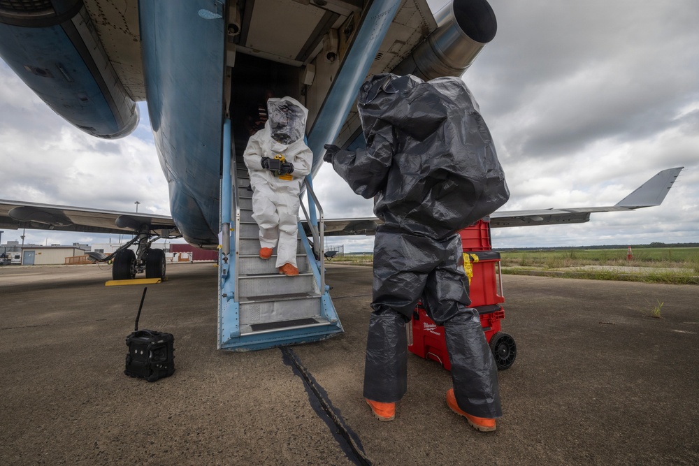 Two individuals wearing full hazmat/chemical protective suits are working around a large aircraft on a tarmac. One person in a white Tyvek-style suit with a clear face shield is descending the aircraft's boarding stairs, while another in a dark gray encapsulated suit stands nearby. A red Milwaukee equipment cart and a black portable device are visible on the tarmac.