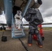 Two individuals wearing full hazmat/chemical protective suits are working around a large aircraft on a tarmac. One person in a white Tyvek-style suit with a clear face shield is descending the aircraft's boarding stairs, while another in a dark gray encapsulated suit stands nearby. A red Milwaukee equipment cart and a black portable device are visible on the tarmac.