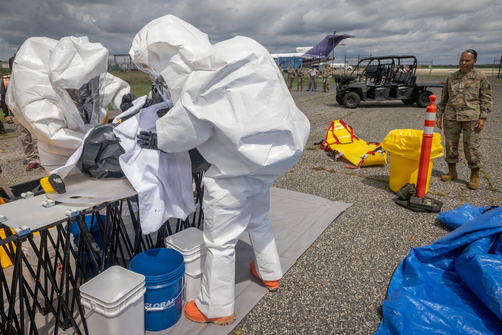 Military personnel are conducting a HAZMAT or CBRN (Chemical, Biological, Radiological, Nuclear) training exercise at what appears to be an airfield or tarmac. Personnel in white full-body protective suits are working at a decontamination station with various containment equipment. A military officer in standard camouflage uniform observes the exercise while additional personnel and vehicles are visible in the background.