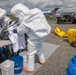 Military personnel are conducting a HAZMAT or CBRN (Chemical, Biological, Radiological, Nuclear) training exercise at what appears to be an airfield or tarmac. Personnel in white full-body protective suits are working at a decontamination station with various containment equipment. A military officer in standard camouflage uniform observes the exercise while additional personnel and vehicles are visible in the background.
