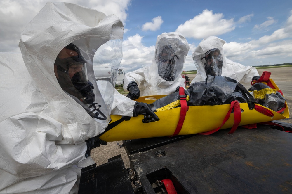 Three personnel wearing white full-body hazmat suits with integrated clear face shields and black rubber gloves are handling a yellow evacuation/decontamination stretcher with red straps on what appears to be an airfield tarmac. A person wrapped in black plastic sheeting is secured on the yellow stretcher, suggesting a simulated contaminated casualty. The setting and equipment indicate a CBRN (Chemical, Biological, Radiological, Nuclear) response drill or training exercise.