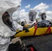 Three personnel wearing white full-body hazmat suits with integrated clear face shields and black rubber gloves are handling a yellow evacuation/decontamination stretcher with red straps on what appears to be an airfield tarmac. A person wrapped in black plastic sheeting is secured on the yellow stretcher, suggesting a simulated contaminated casualty. The setting and equipment indicate a CBRN (Chemical, Biological, Radiological, Nuclear) response drill or training exercise.