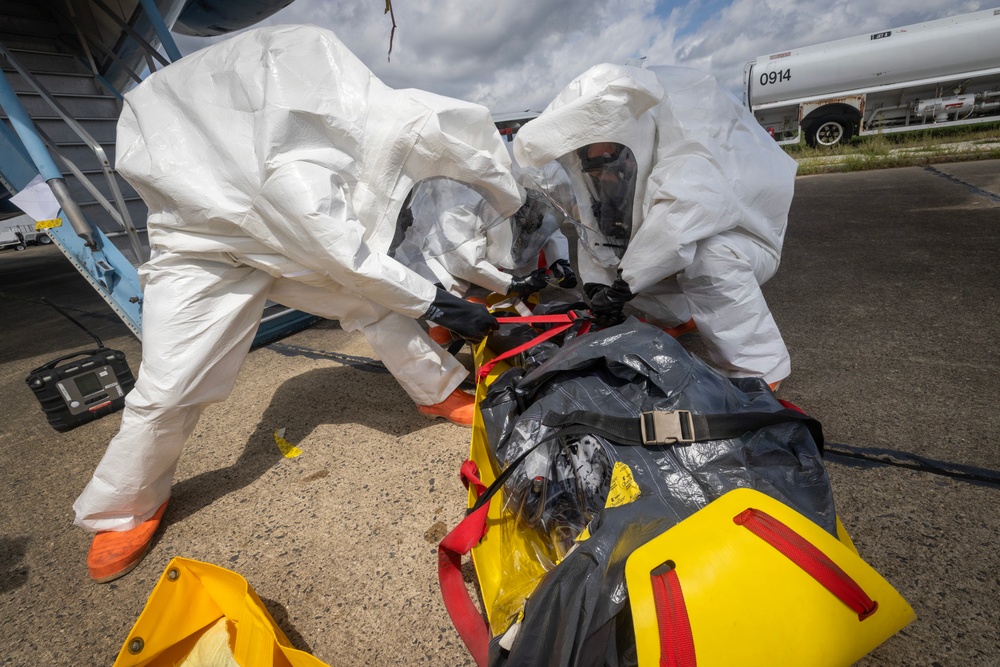 Two personnel in white Tyvek hazmat suits and full-face respirator masks are handling a patient secured in a yellow CBRN/hazmat evacuation stretcher with a dark protective covering. The scene appears to be an outdoor tarmac or airfield environment, suggesting a hazardous materials medical response or decontamination exercise. A gas detection device is visible on the ground nearby, and a tanker truck is visible in the background.