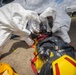 Two personnel in white Tyvek hazmat suits and full-face respirator masks are handling a patient secured in a yellow CBRN/hazmat evacuation stretcher with a dark protective covering. The scene appears to be an outdoor tarmac or airfield environment, suggesting a hazardous materials medical response or decontamination exercise. A gas detection device is visible on the ground nearby, and a tanker truck is visible in the background.