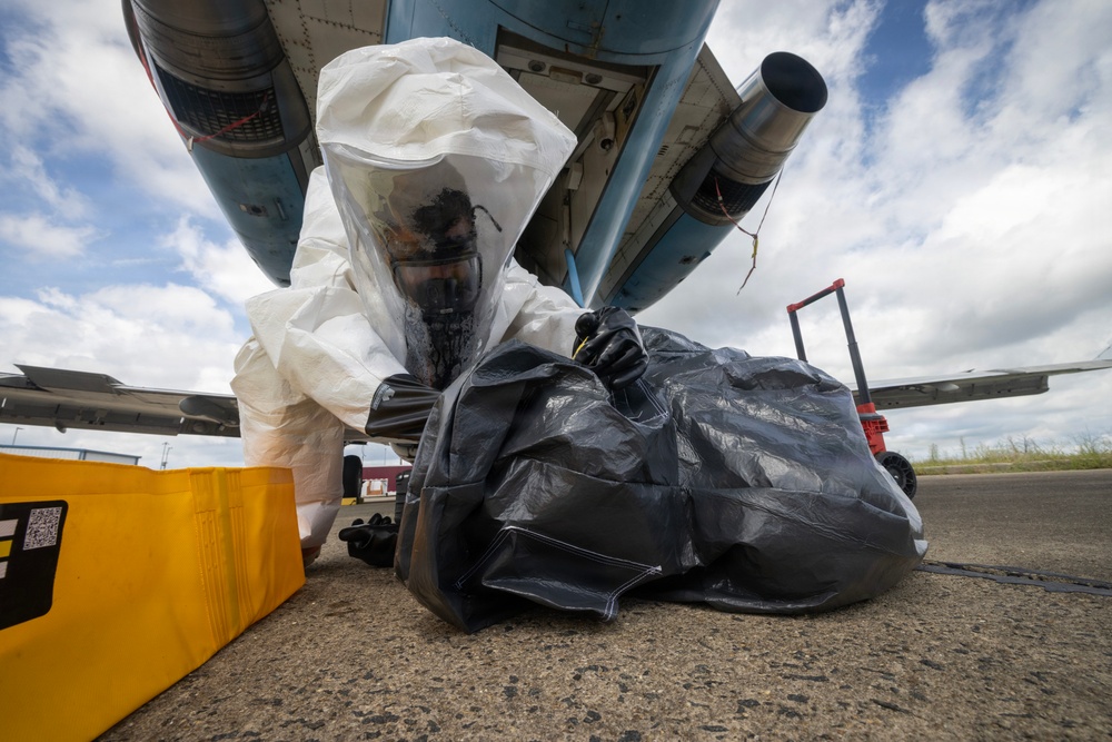 A person wearing a full white hazmat suit with an integrated respirator mask is handling a large black plastic bag on an airport tarmac beneath the fuselage of an aircraft. The scene appears to depict a hazardous materials response or decontamination procedure. A yellow containment or equipment case is visible to the left of the subject.