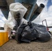 A person wearing a full white hazmat suit with an integrated respirator mask is handling a large black plastic bag on an airport tarmac beneath the fuselage of an aircraft. The scene appears to depict a hazardous materials response or decontamination procedure. A yellow containment or equipment case is visible to the left of the subject.