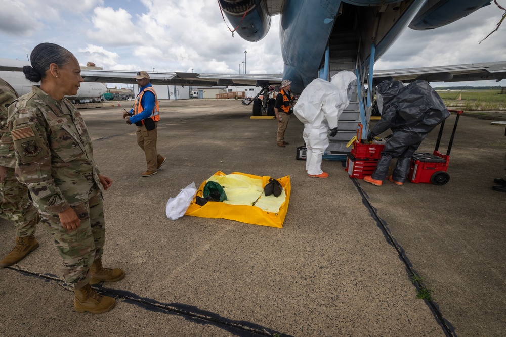 A military or emergency response exercise is taking place on an airfield tarmac next to a large aircraft. Personnel in hazmat suits are working near the aircraft stairs alongside individuals in orange safety vests, while a uniformed military service member observes. A yellow containment tray with absorbent materials is visible on the ground, suggesting a hazardous materials response scenario.