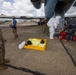 A military or emergency response exercise is taking place on an airfield tarmac next to a large aircraft. Personnel in hazmat suits are working near the aircraft stairs alongside individuals in orange safety vests, while a uniformed military service member observes. A yellow containment tray with absorbent materials is visible on the ground, suggesting a hazardous materials response scenario.