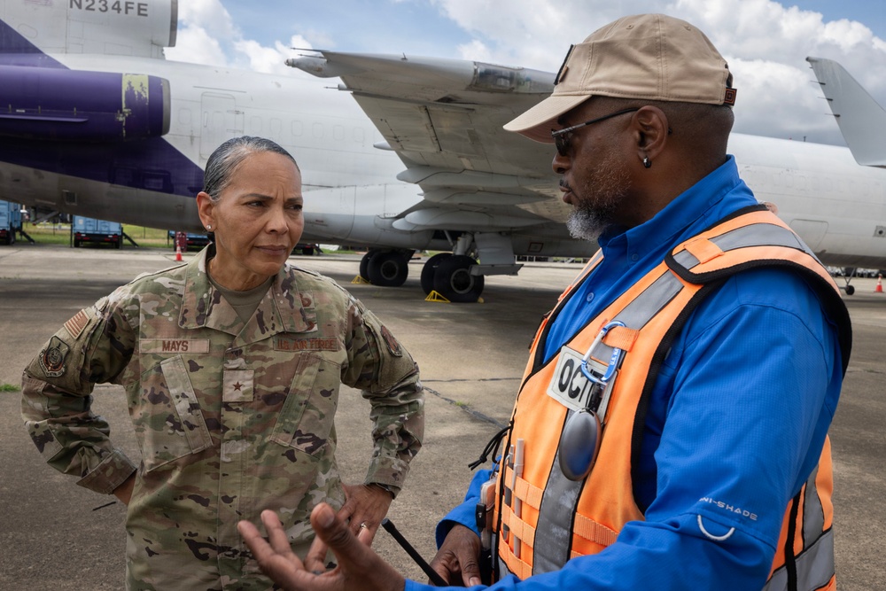 A U.S. Air Force officer with the name tape 'MAYS' and a one-star general rank insignia (Brigadier General) is engaged in conversation with a civilian ground operations worker wearing an orange high-visibility safety vest on an airport tarmac. Large aircraft are visible in the background, including one with a purple livery bearing the registration 'N234FE'. The setting appears to be a military or joint-use airfield during daylight hours.