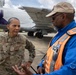 A U.S. Air Force officer with the name tape 'MAYS' and a one-star general rank insignia (Brigadier General) is engaged in conversation with a civilian ground operations worker wearing an orange high-visibility safety vest on an airport tarmac. Large aircraft are visible in the background, including one with a purple livery bearing the registration 'N234FE'. The setting appears to be a military or joint-use airfield during daylight hours.