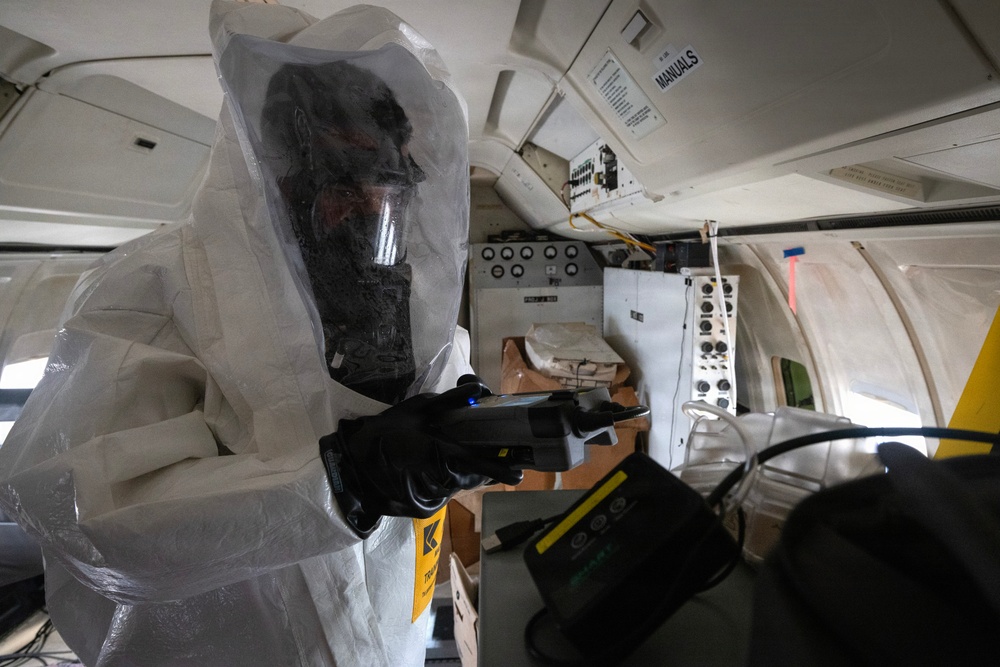 A person wearing a full white hazmat/Tyvek protective suit with a black respirator/gas mask is operating inside what appears to be the interior of a small aircraft or similar vehicle. They are holding a handheld scanning or detection device. The environment contains equipment panels, cardboard boxes, and various instruments consistent with a decontamination or hazardous materials assessment operation.