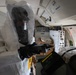 A person wearing a full white hazmat/Tyvek protective suit with a black respirator/gas mask is operating inside what appears to be the interior of a small aircraft or similar vehicle. They are holding a handheld scanning or detection device. The environment contains equipment panels, cardboard boxes, and various instruments consistent with a decontamination or hazardous materials assessment operation.