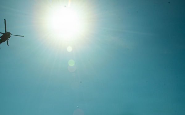 A military scene captured in silhouette showing soldiers on the ground observing or communicating with a helicopter flying overhead. The image is taken from a low angle looking upward into a bright sun, creating strong backlit silhouettes. One soldier appears to be signaling or saluting toward the aircraft while another holds what appears to be a radio.