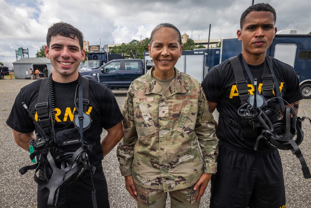 Three military personnel pose together outdoors in what appears to be a military installation or training area. The center figure wears a U.S. Air Force OCP (Operational Camouflage Pattern) uniform with the name tape 'MAYS' and a single star insignia indicating a Brigadier General rank. The two flanking individuals wear black Army PT (Physical Training) shirts and are equipped with what appear to be scuba or breathing apparatus harnesses.